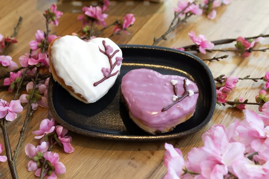 Two heart-shaped donuts decorated with cherry blossom branches sit on a table staged with cherry blossoms. 